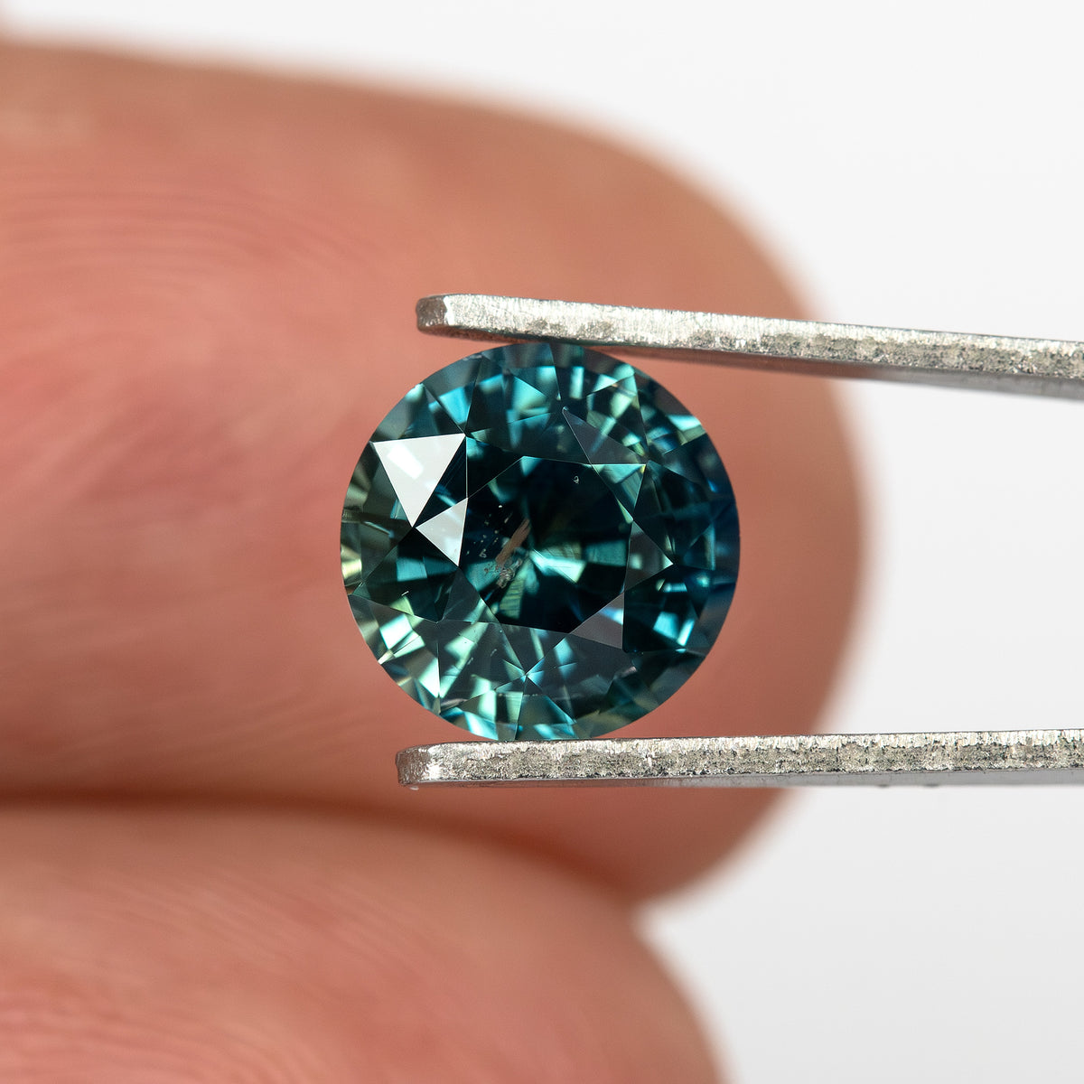 Round deep teal blue-green sapphire held in tweezers, viewed from the side against a fingertip background.