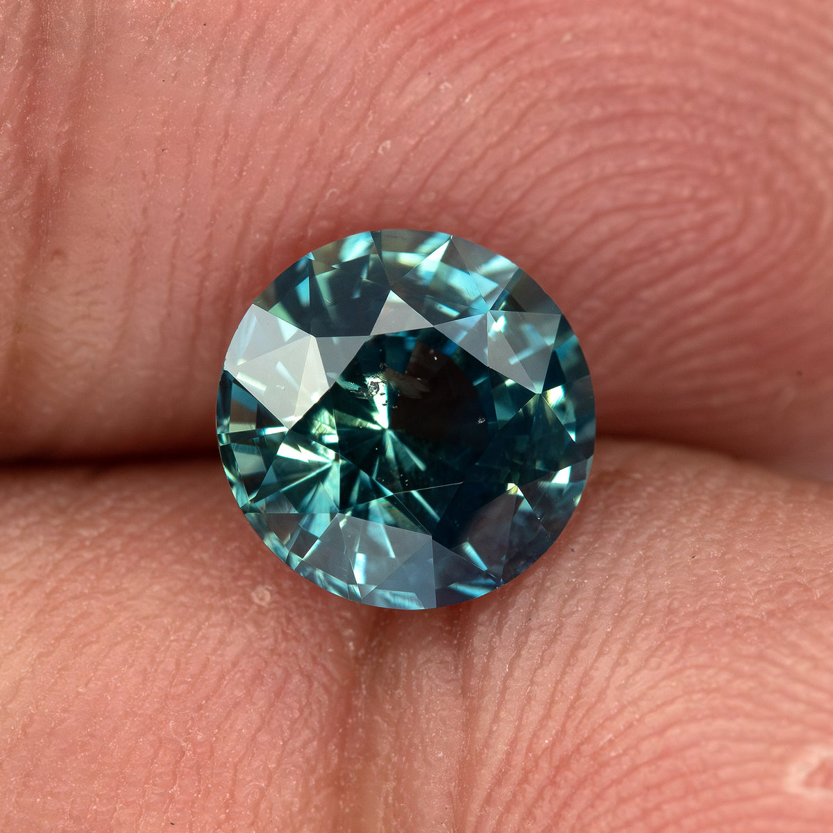 Round deep teal blue-green sapphire on fingertips, viewed face-up showing facets and vivid color.
