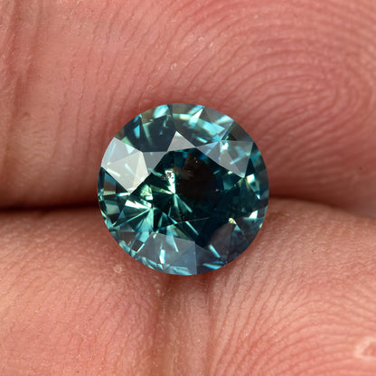 Round deep teal blue-green sapphire on fingertips, viewed face-up showing facets and vivid color.