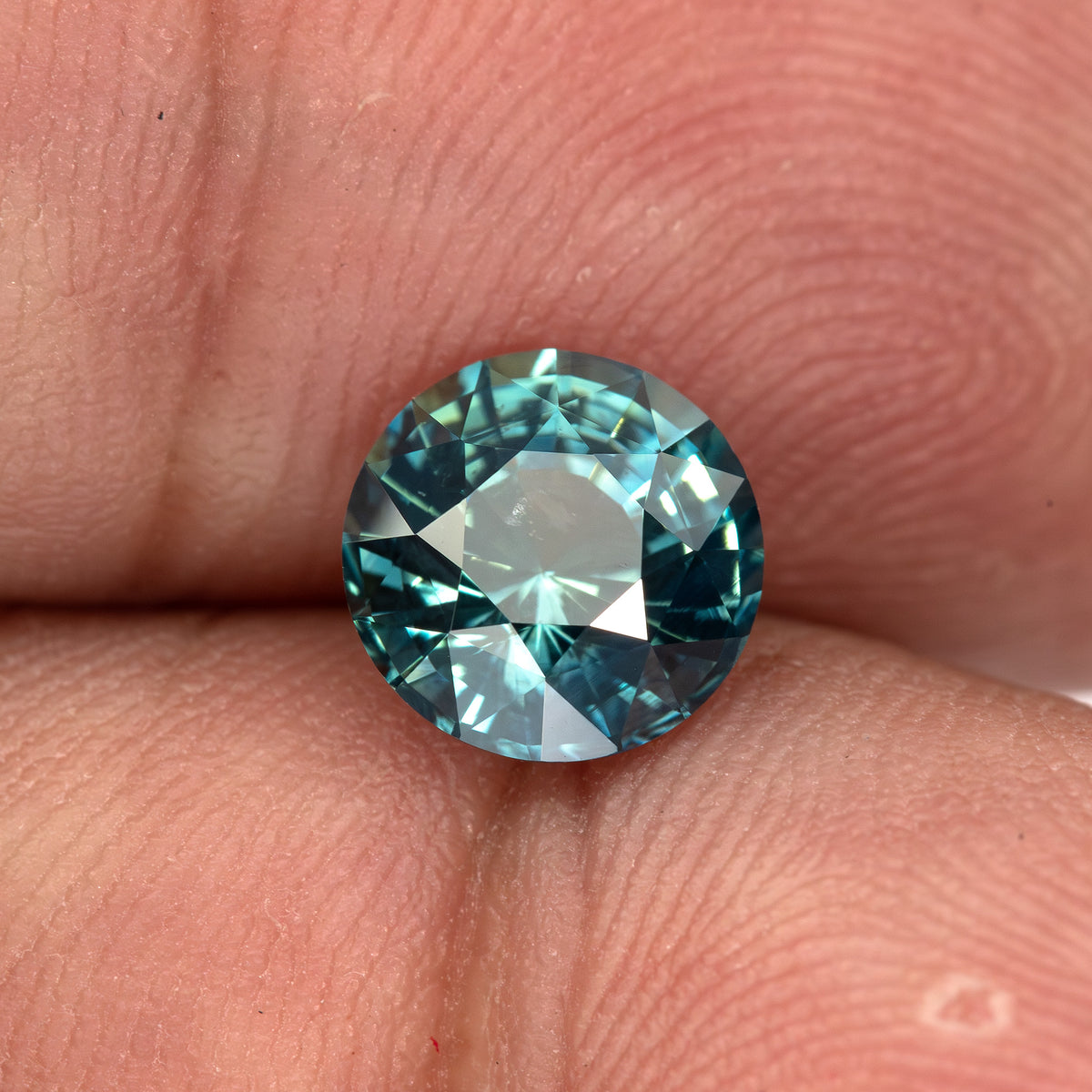 Round deep teal blue-green sapphire shown face-up on fingertips.