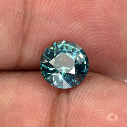 Round deep teal blue-green sapphire shown face-up on fingertips.