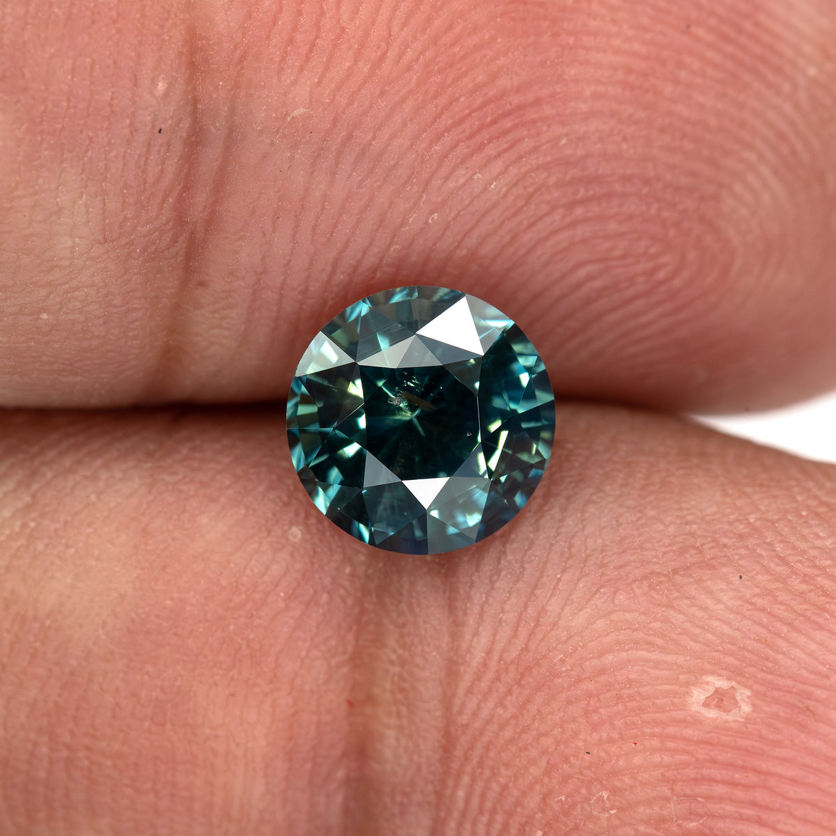 Round deep teal blue-green sapphire resting on fingertips, showcasing its face-up view.