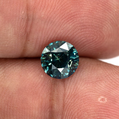 Round deep teal blue-green sapphire resting on fingertips, showcasing its face-up view.