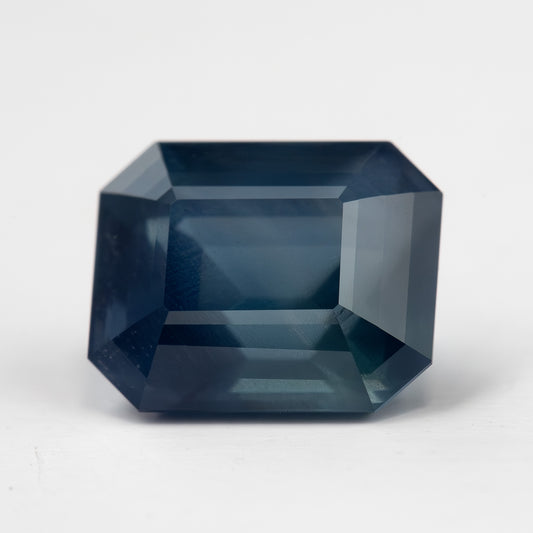 Face-up view of a deep teal-blue emerald-cut sapphire on white,, showing clipped corners and broad step facets.