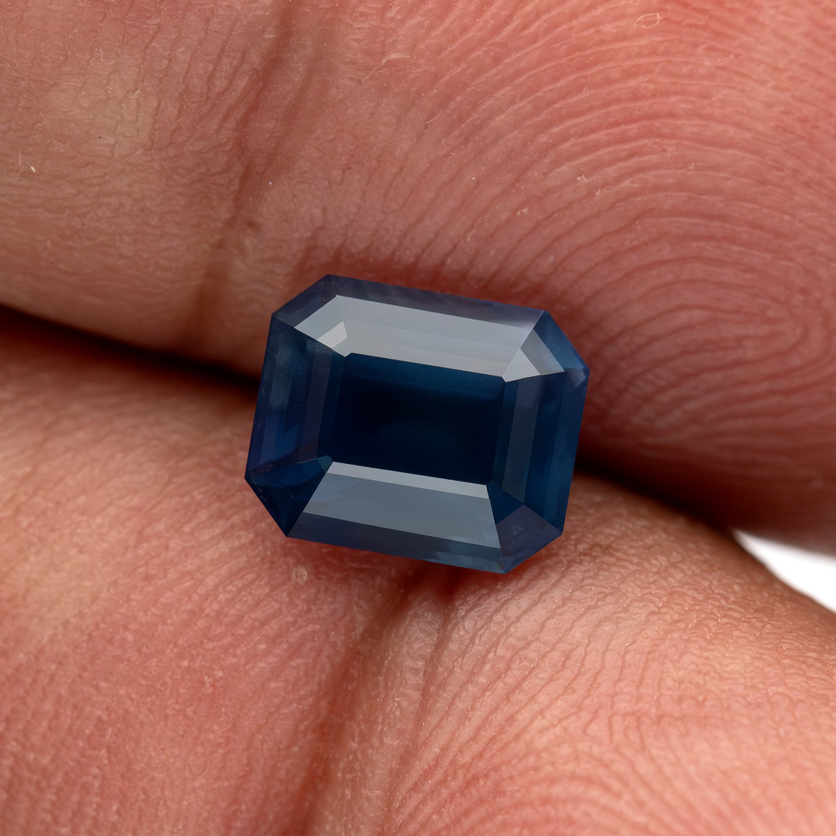 Zafiro de frente equilibrado sobre las puntas de los dedos para escala, con su centro azul profundo y facetas escalonadas superiores brillantes visibles.