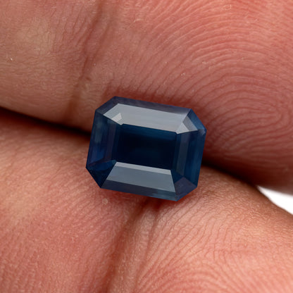 Zafiro de frente equilibrado sobre las puntas de los dedos para escala, con su centro azul profundo y facetas escalonadas superiores brillantes visibles.