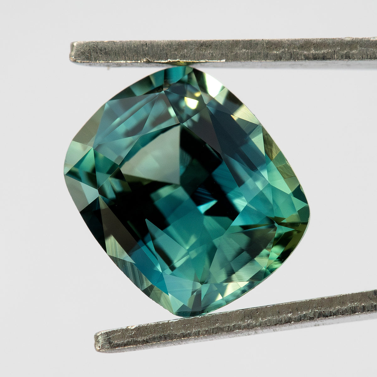 Face-up teal sapphire held between tweezers against a pale background, showing its cushion outline and light and dark blue-green reflections.
