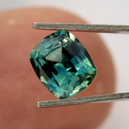 Face-up sapphire held between tweezers in front of a fingertip, showing its cushion outline, teal reflections and approximate scale.