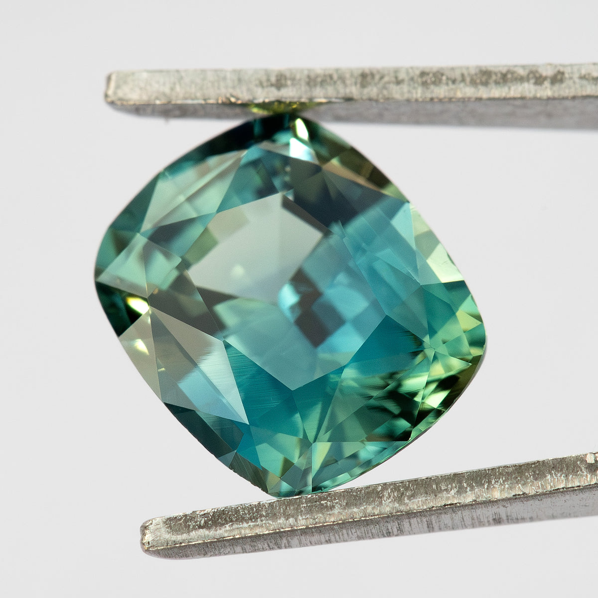 Alternate face-up view of the cushion sapphire between tweezers, showing pale green and blue reflections across its facets.