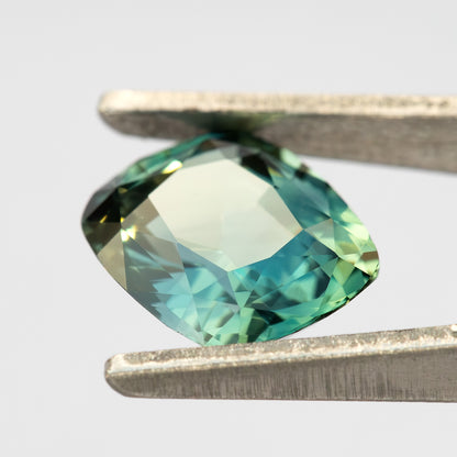 Tilted face-up view of the sapphire between tweezers, showing its cushion outline and a broad pale reflection across the table.