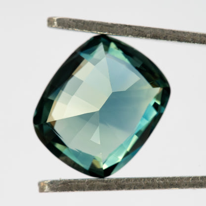 Pavilion-facing view of the sapphire between tweezers against a pale background, showing broad green-blue back facets.
