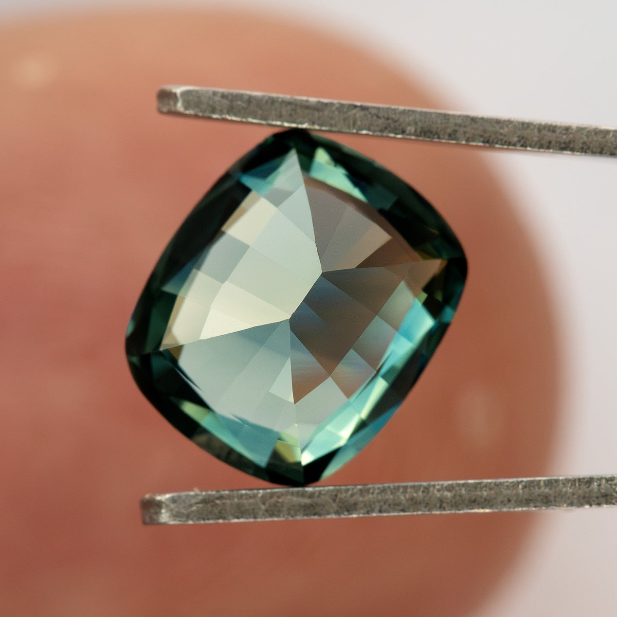 Pavilion-facing sapphire between tweezers in front of a fingertip, showing broad back facets and green-blue reflections.