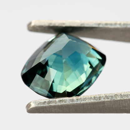 Angled pavilion view of the sapphire between tweezers, showing broad back facets and pale blue-green reflections.