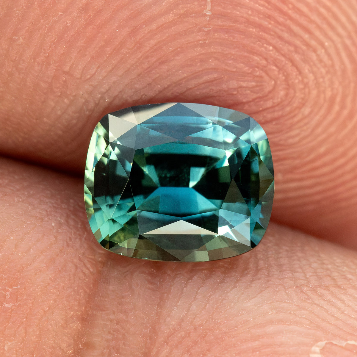 Cushion sapphire resting on a fingertip for scale, with teal facets and a dark horizontal reflection across the center.