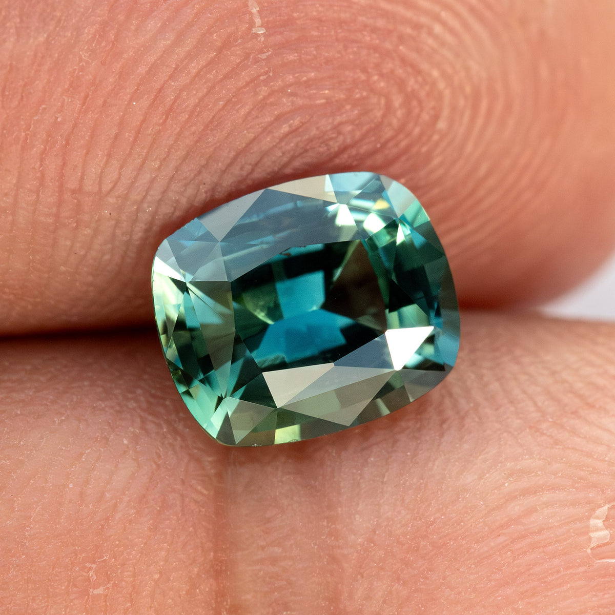Slightly rotated face-up view of the sapphire on a fingertip, showing approximate scale and contrasting teal reflections.