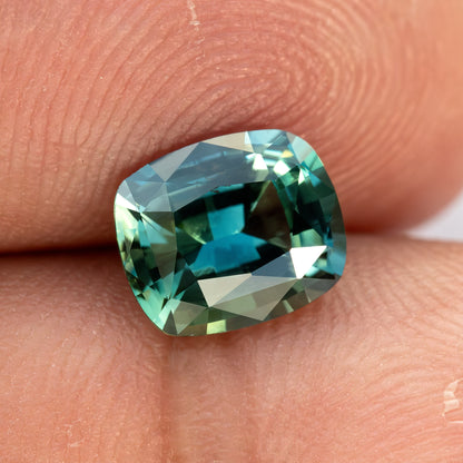 Slightly rotated face-up view of the sapphire on a fingertip, showing approximate scale and contrasting teal reflections.