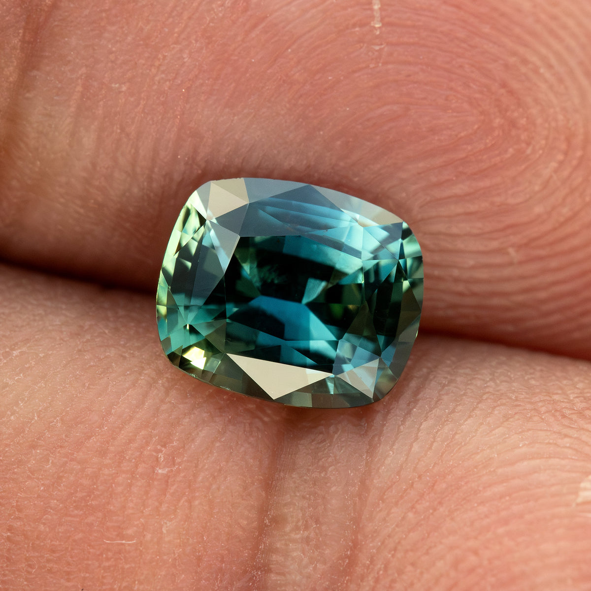 Angled face-up view of the sapphire resting on a fingertip, showing its cushion proportions and blue-green reflections.