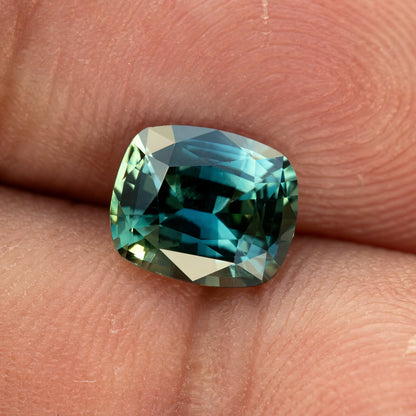 Angled face-up view of the sapphire resting on a fingertip, showing its cushion proportions and blue-green reflections.