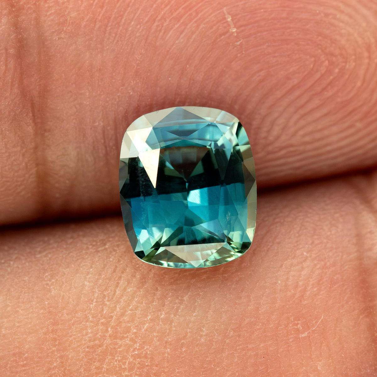 Sapphire resting lengthwise on a fingertip, showing its cushion outline and contrasting dark and bright blue-green reflections.