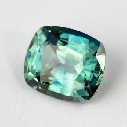 Angled face-up view of the loose sapphire on a pale surface, showing rounded cushion corners and pale green-blue reflections.