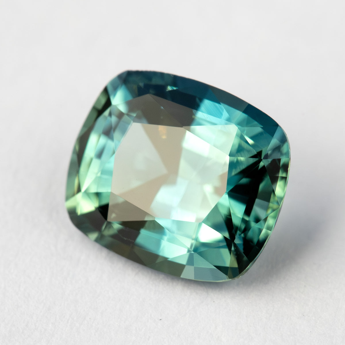 Angled face-up sapphire on a pale surface, with a broad light reflection across the table and darker teal edges.