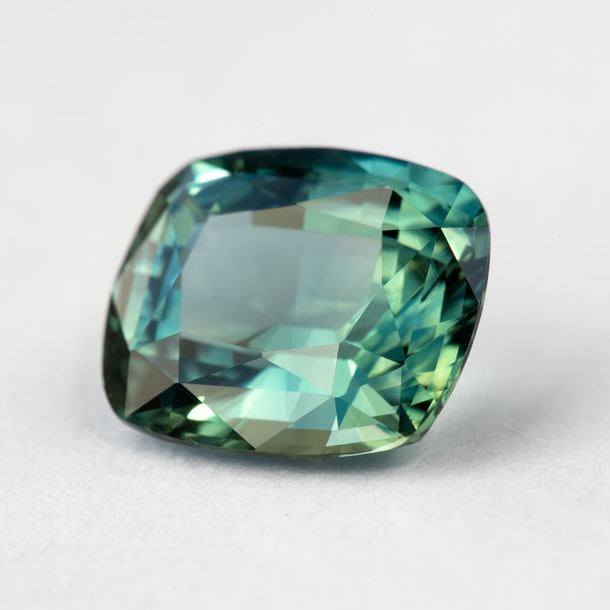 Low oblique face-up view of the cushion sapphire on a pale surface, showing the crown and light green-blue reflections.