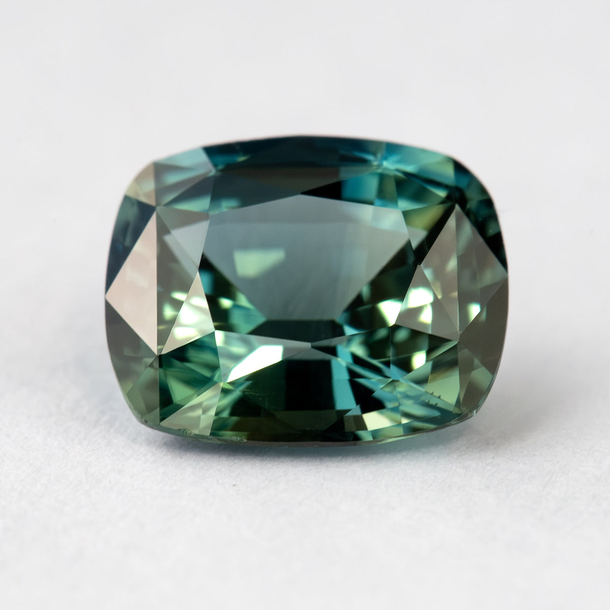 Face-up view of the sapphire on a pale surface, with its longer dimension horizontal and contrasting light and dark teal reflections.