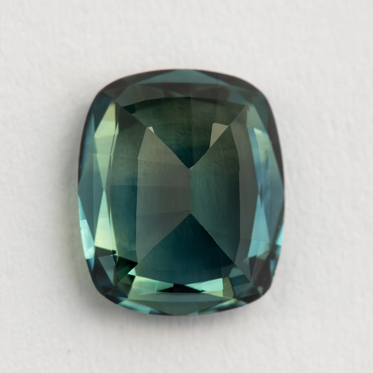 Pavilion-facing view of the sapphire on a pale surface, with its longer dimension vertical and dark green-blue back facets visible.