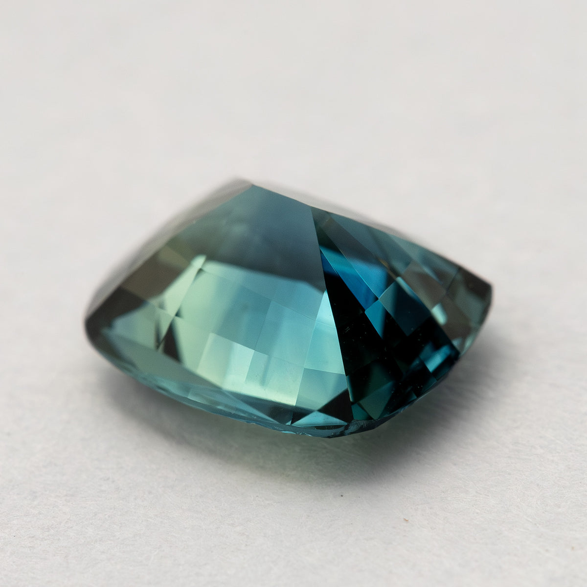 Oblique view of the sapphire resting pavilion-up on a pale surface, showing its tapered back and teal pavilion facets.