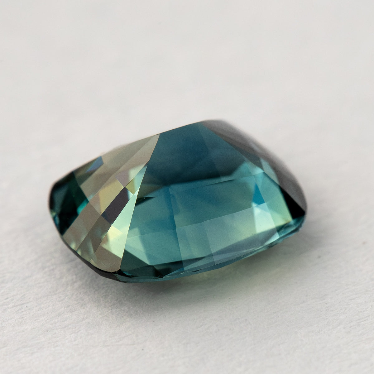 Three-quarter pavilion-up view of the sapphire on a pale surface, showing its tapered profile and broad blue-green reflections.