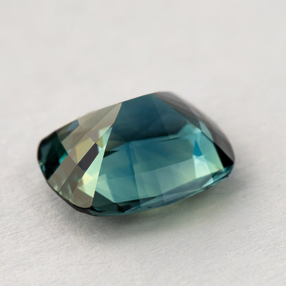 Three-quarter pavilion-up view of the sapphire on a pale surface, showing its tapered profile and broad blue-green reflections.