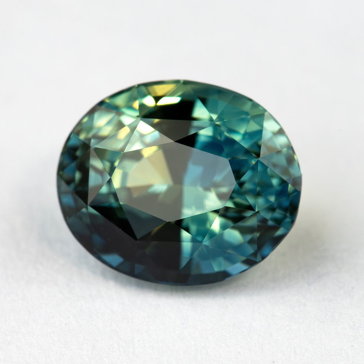Close face-up view of the oval teal blue-green sapphire on a pale background, showing its mixed facet pattern and contrasting reflections.
