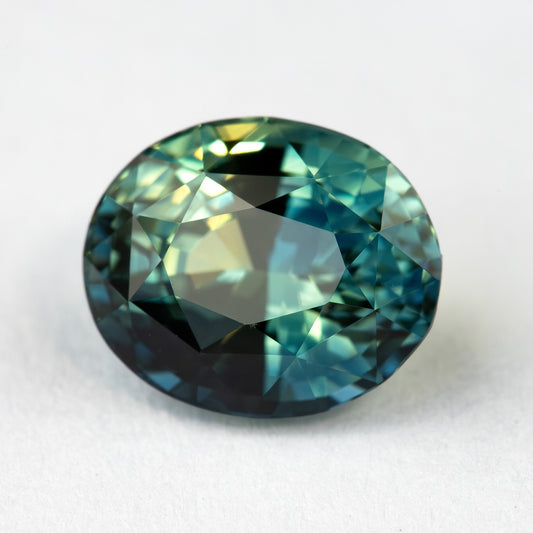 Close face-up view of the oval teal blue-green sapphire on a pale background, showing its mixed facet pattern and contrasting reflections.