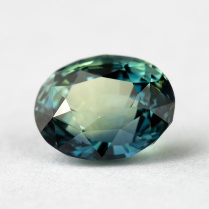 Angled face-up view of the sapphire on a pale surface, showing pale green and blue reflections across the oval.