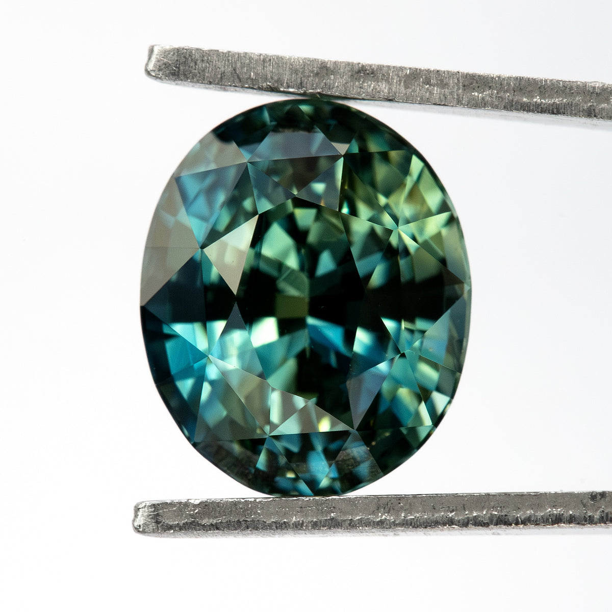 Face-up oval sapphire held vertically between tweezers, showing the outline, deep teal reflections and facet pattern.