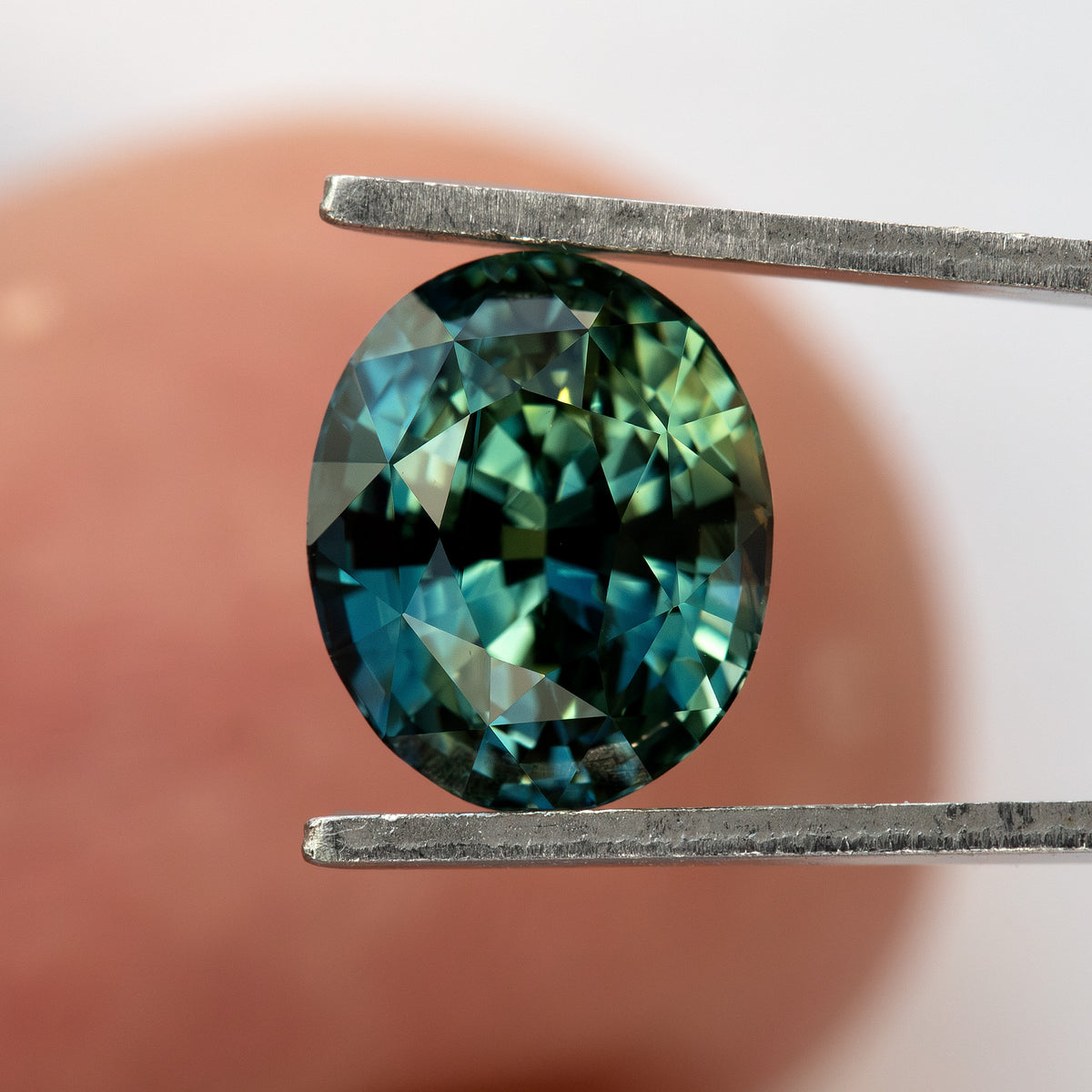 Face-up oval sapphire between tweezers in front of a fingertip, showing approximate scale and deep blue-green reflections.