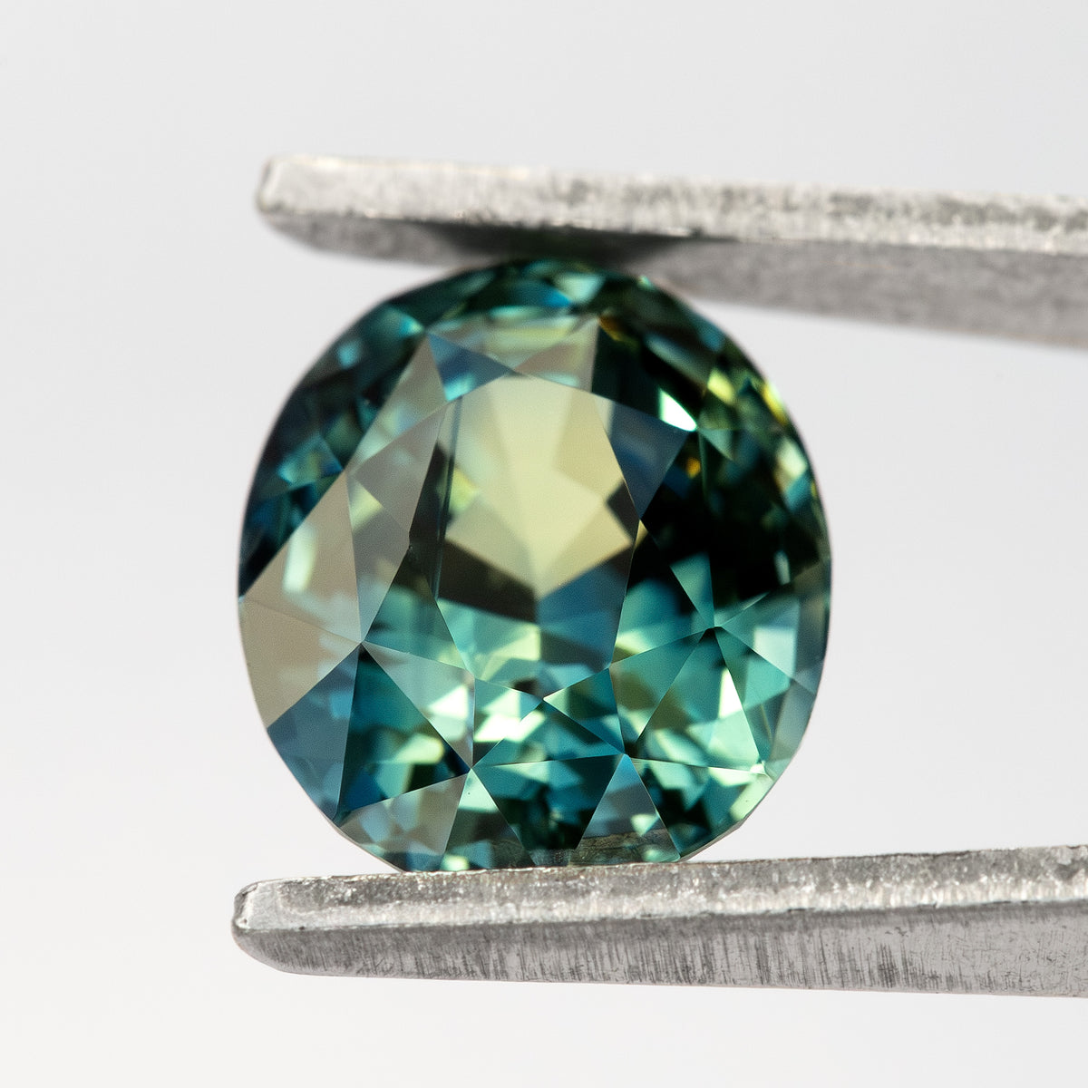 Face-up oval sapphire held between tweezers against a pale background, showing its mixed cut and blue-green color.