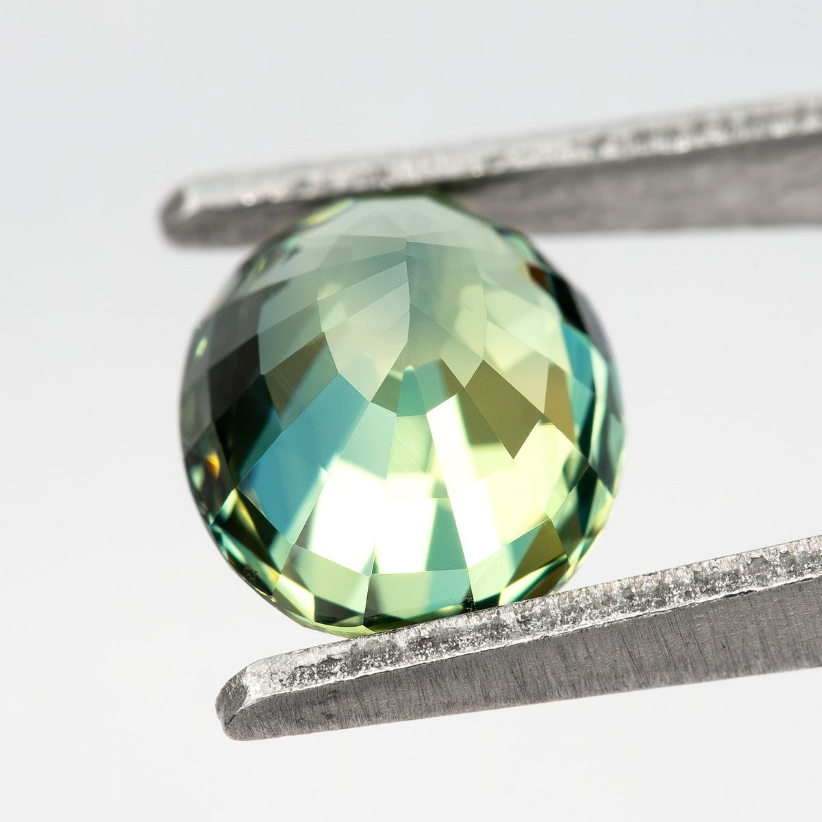 Pavilion-facing oval sapphire between tweezers, showing the back facet pattern and pale green-blue reflections.