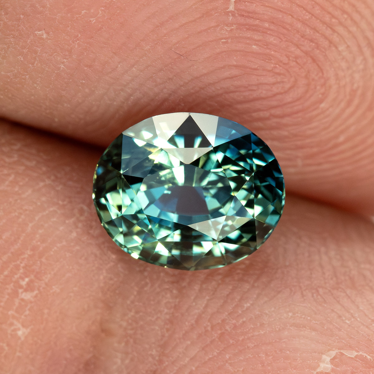 Face-up teal sapphire resting on a fingertip for scale, showing its oval proportions and blue-green reflections.