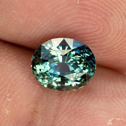 Face-up teal sapphire resting on a fingertip for scale, showing its oval proportions and blue-green reflections.