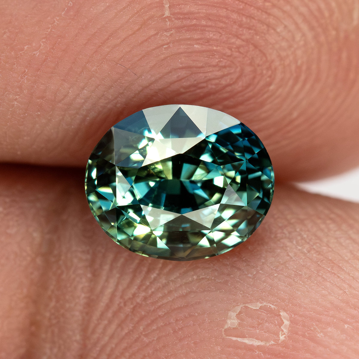 Face-up sapphire on a fingertip, showing its oval outline, approximate scale and bright teal reflections.