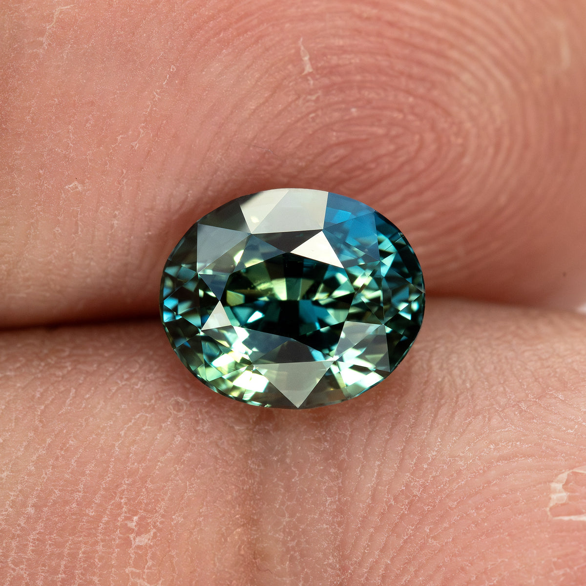 Alternate fingertip scale view of the face-up sapphire, showing its oval outline and cooler blue-green reflections.