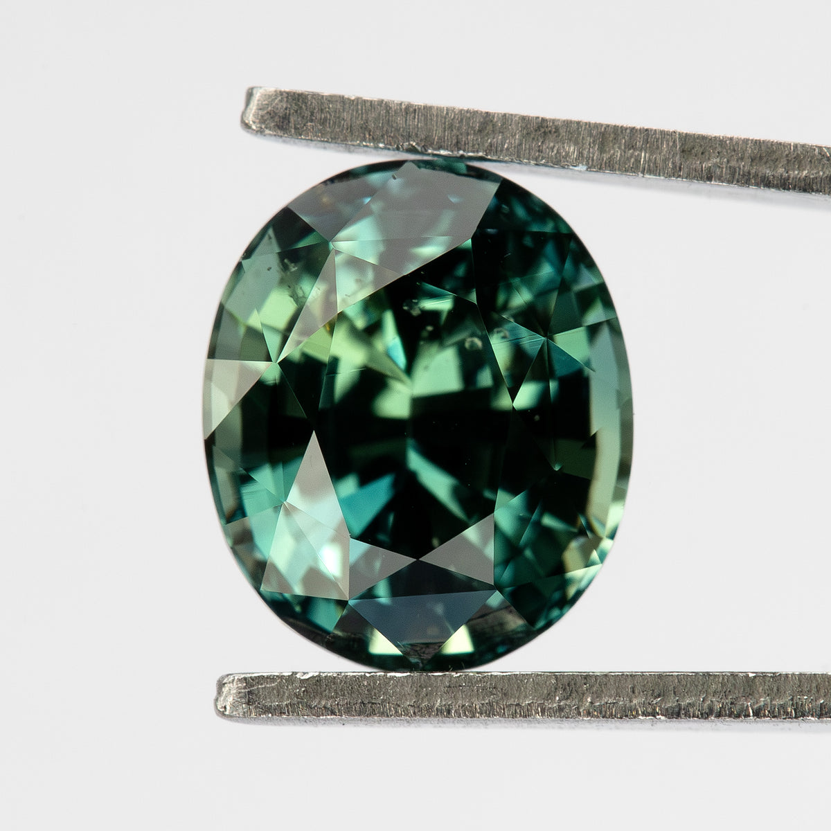 Oval-shaped teal blue-green sapphire viewed face-up between tweezers.