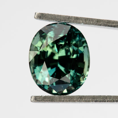 Oval-shaped teal blue-green sapphire viewed face-up between tweezers.