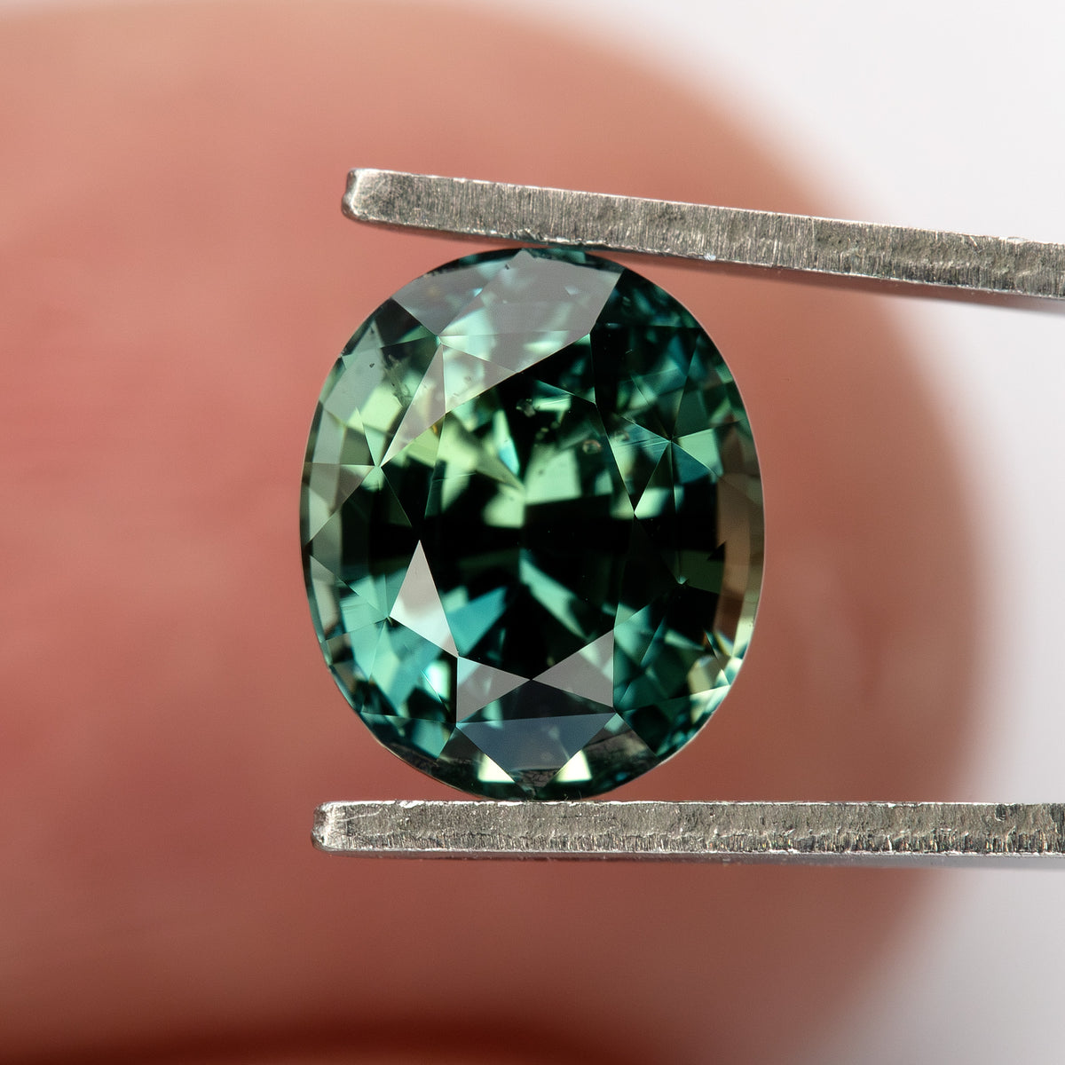 Oval teal blue-green sapphire held by tweezers, face-up view with a fingertip background.