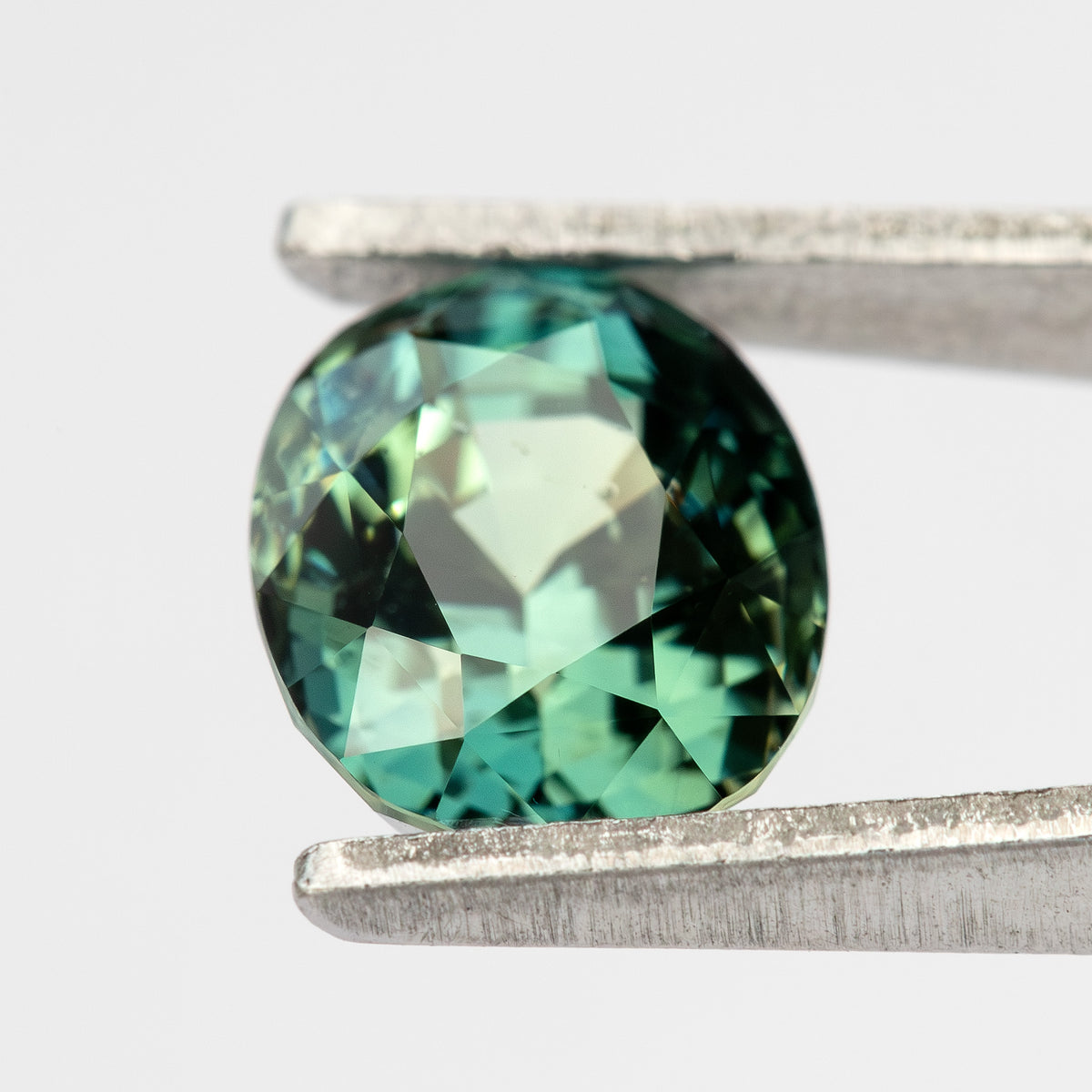 Oval-shaped teal blue-green sapphire held with tweezers, showcasing its facets from a top-down angle.