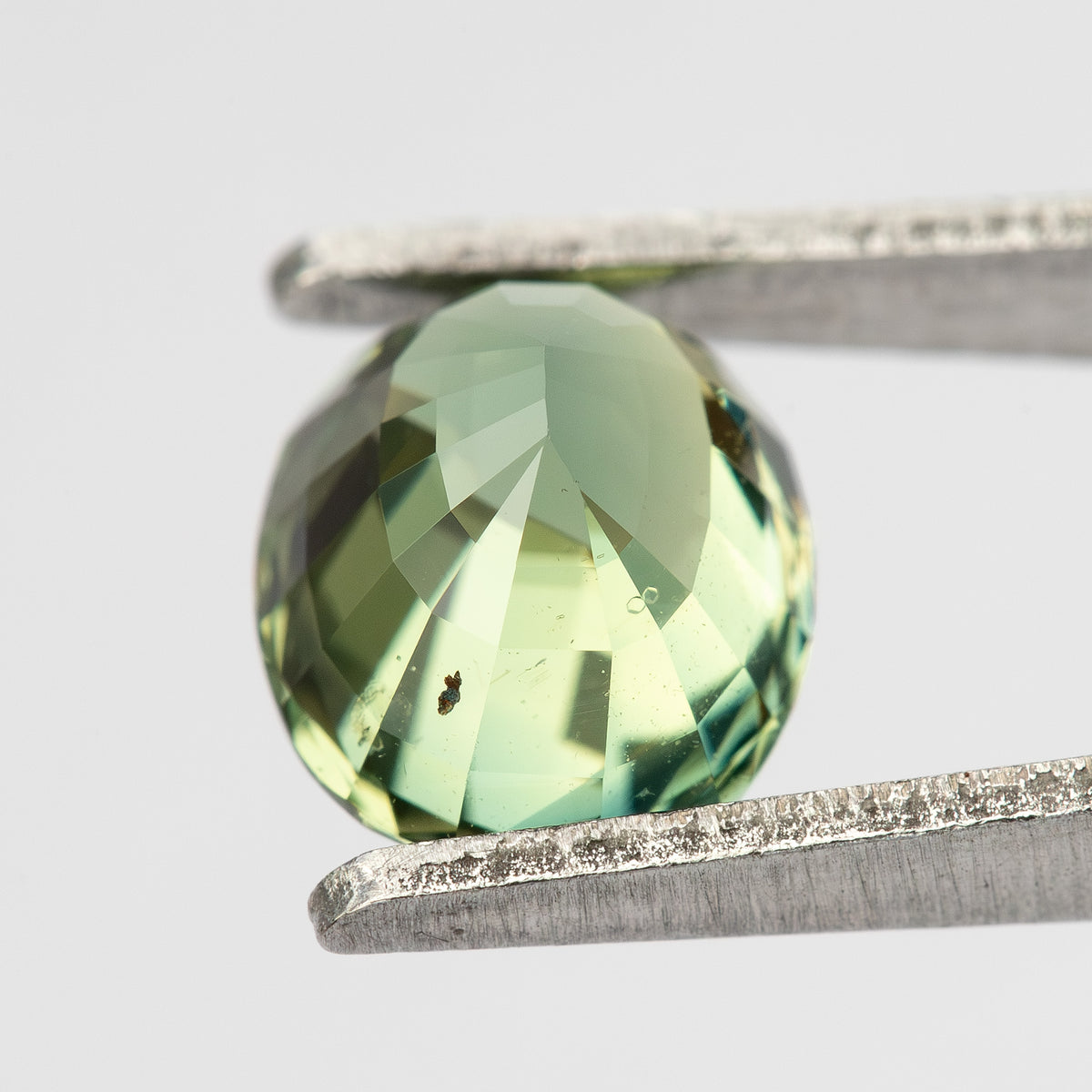 Oval mixed-cut teal blue-green sapphire held in tweezers, showing the pavilion view with faceted reflections.