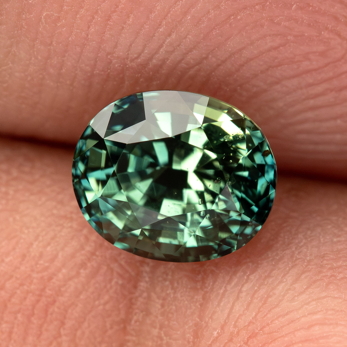 Oval-cut teal blue-green sapphire shown face-up between fingertips.