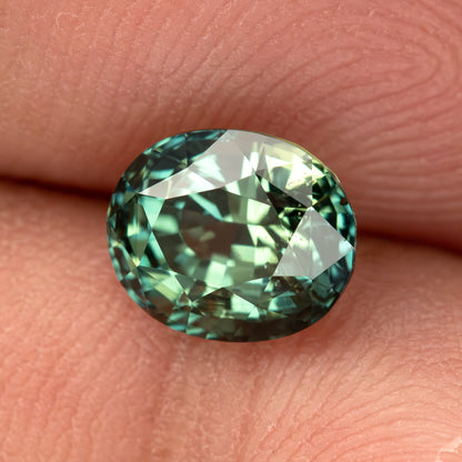 Oval-shaped teal blue-green sapphire displayed face-up on fingers.