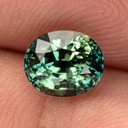 Oval teal blue-green sapphire shown face-up on a fingertip, highlighting its facets and color variations.